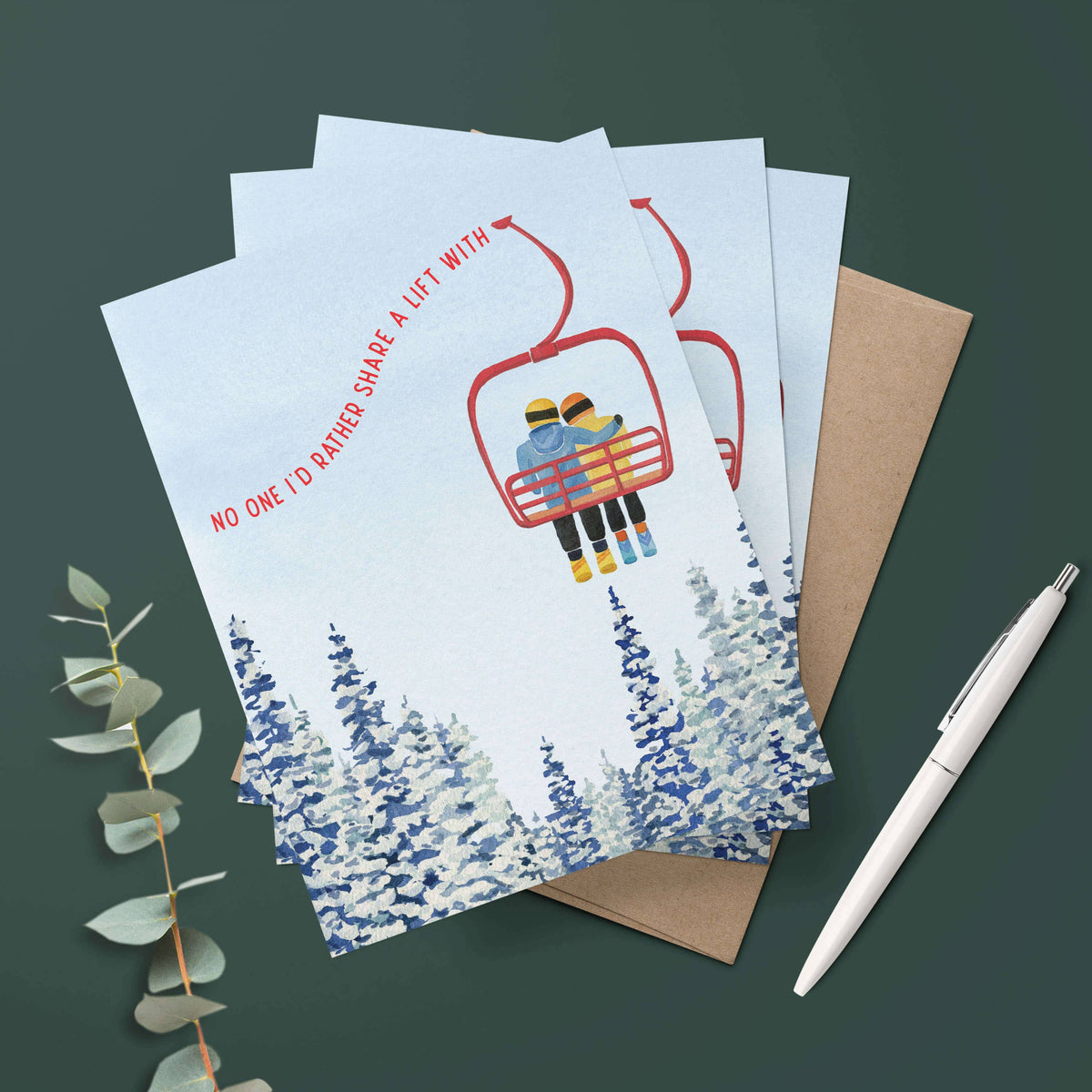 Three Chairlift Love Cards fanned out on a dark green surface with kraft envelopes and a white pen, styled with eucalyptus leaves