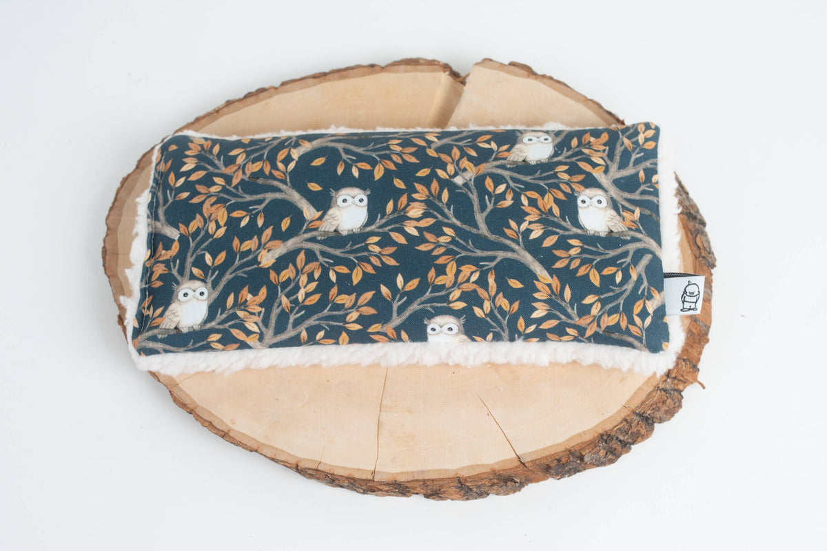 Night Owl Deep Rest eye pillow in a dark teal fabric of owls on leafy branches, cream sherpa edges, on a wood slice