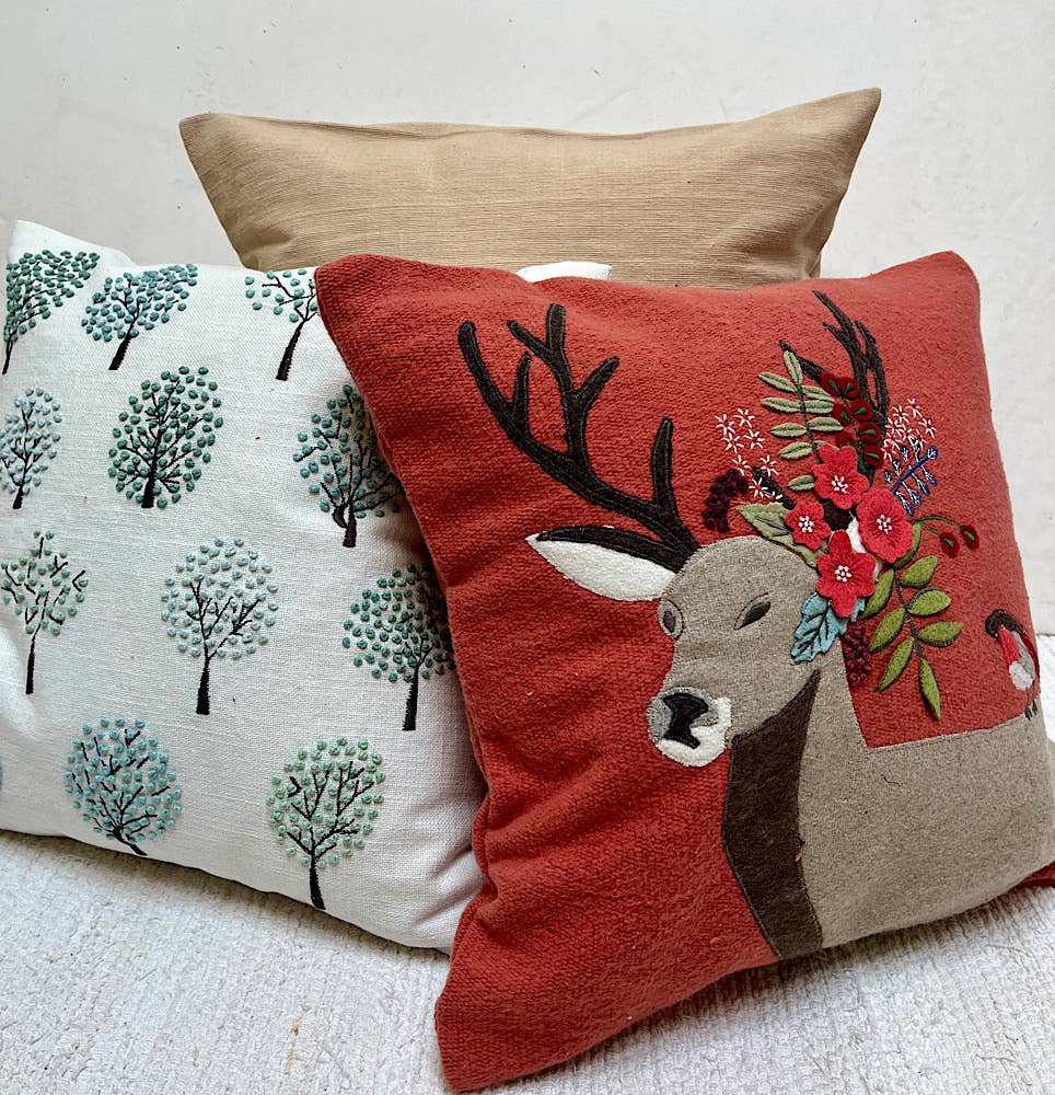 Rust red pillow with an appliqued deer and flowers stacked in front of a white tree-embroidered pillow and a tan pillow