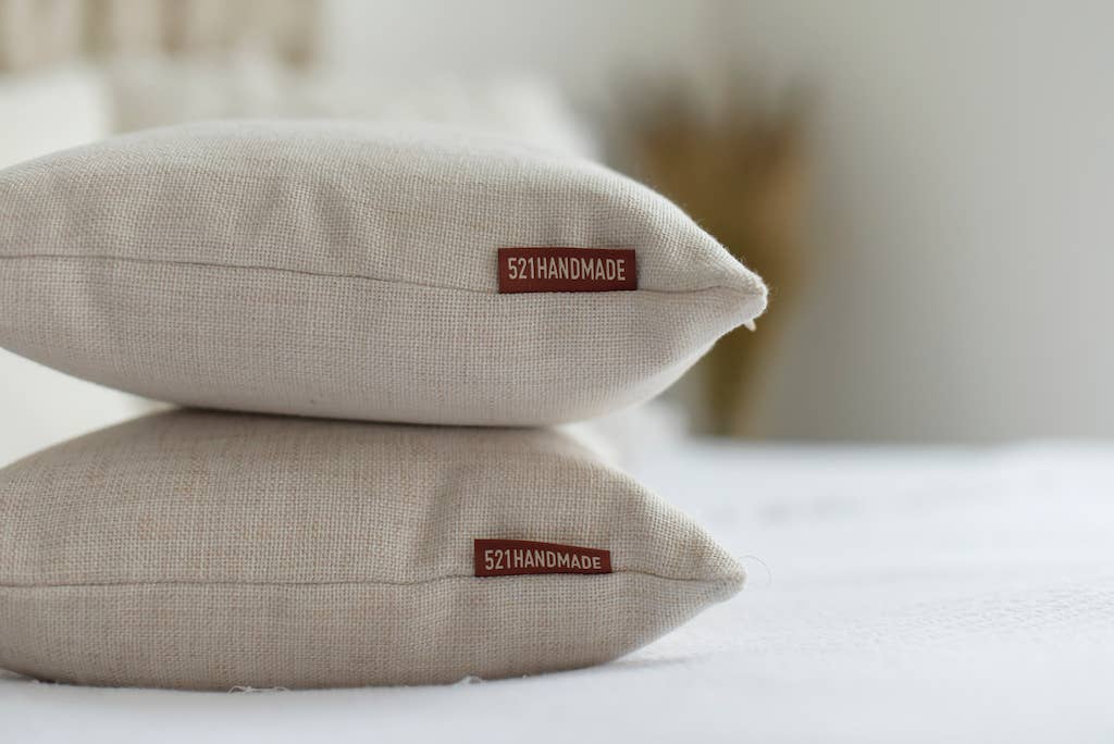 Close-up of two stacked natural linen lumbar pillows with visible seams and the red 521handmade fabric tag on a dark table