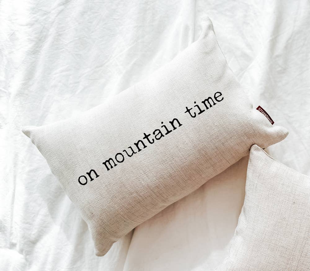 On Mountain Time Linen Pillow lying on a white bed next to a plain natural linen pillow, alternate angle