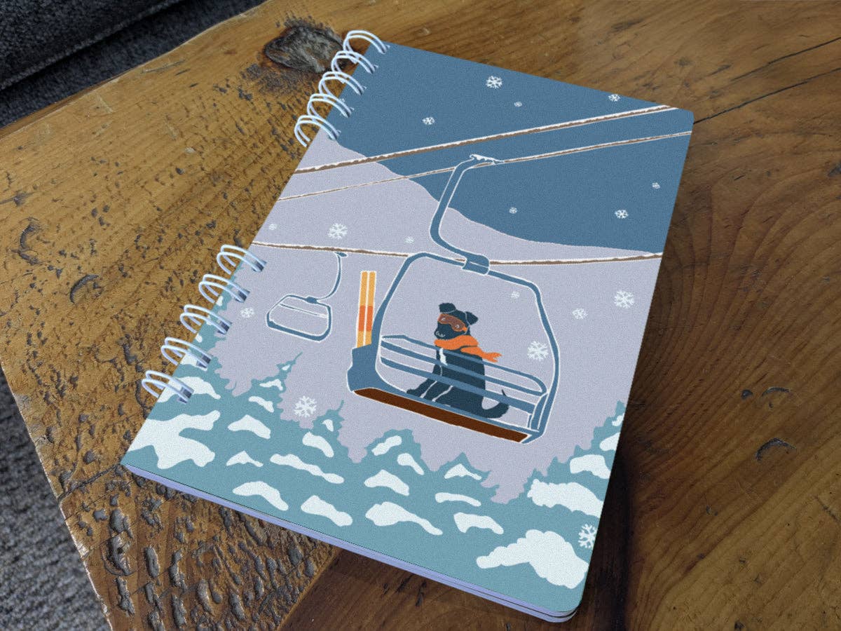 Ski Lift Pup spiral notebook resting on a wooden table, its cover showing a pup in an orange scarf and goggles riding a chairlift above snowy pines