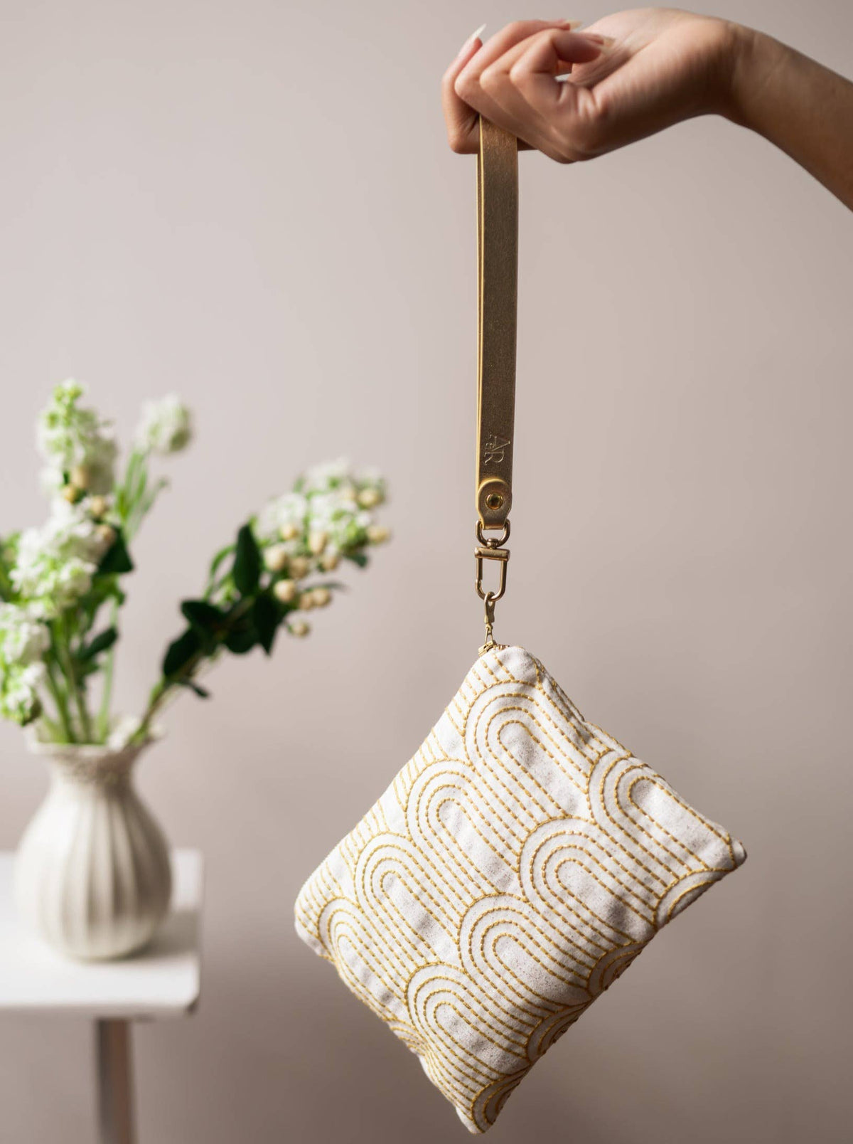 A woman holding a Josephine Clutch wristlet purse by its gold strap, cream fabric with gold Art Deco arch embroidery