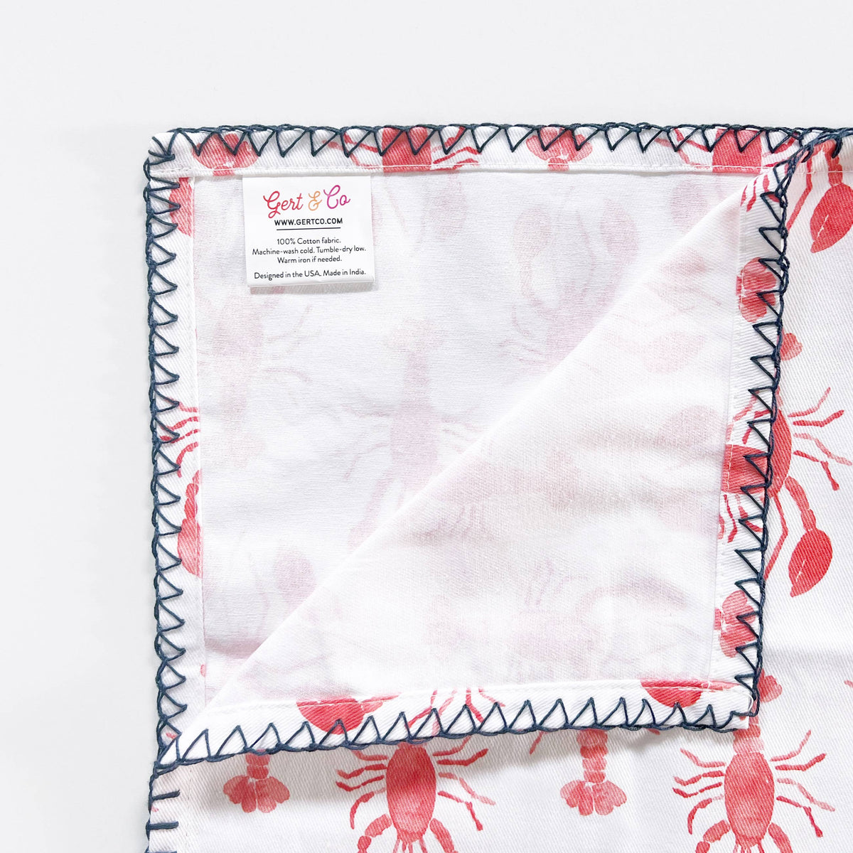 Folded-back corner of a lobster napkin showing the Gert & Co care label and the navy zigzag stitching