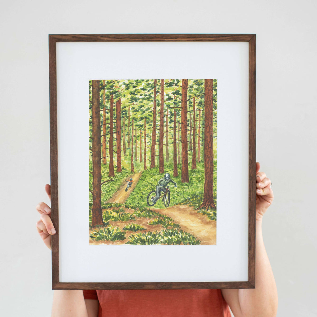 Framed Pedaling Through the Pines Watercolor Art Print in a dark wood frame with white mat, hanging on a wood paneled cabin wall above a bed with white linens and an orange pillow