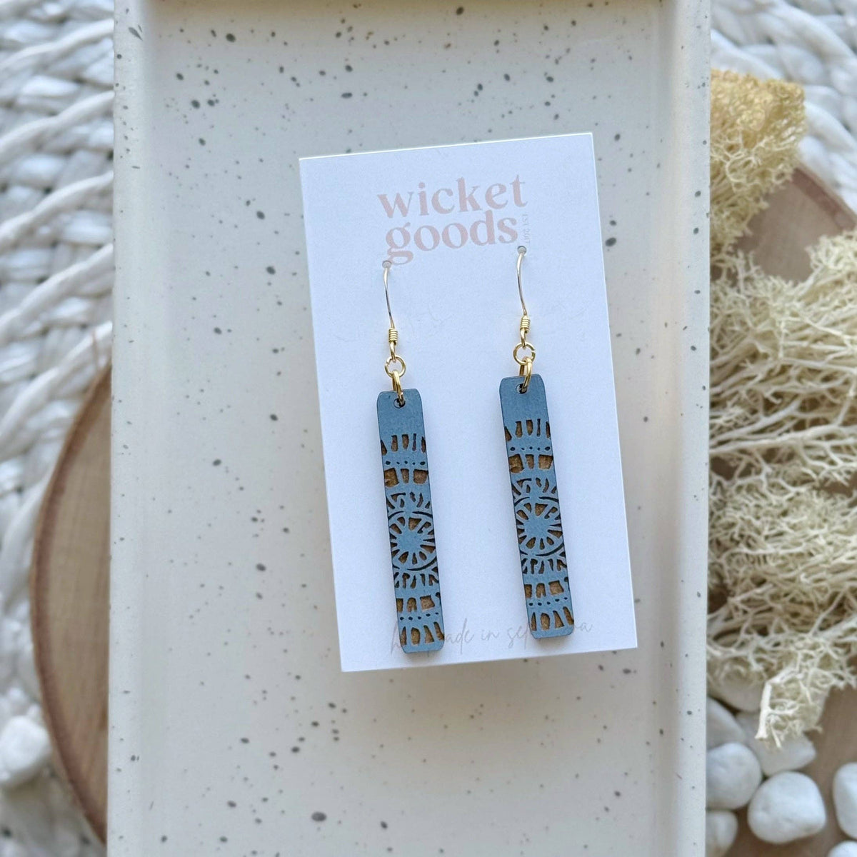 A pair of rectangular drop earrings with a dusty blue laser-cut geometric pattern and gold fishhook wires, displayed on a white 'wicket goods' card. The card is set on a speckled tray with dried botanicals and white stones in the background.