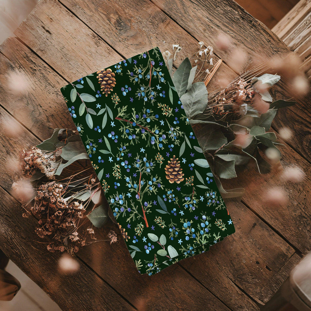 Winter Juniper Tea Towel folded on a rustic wood table surrounded by dried eucalyptus and pinecones, showing juniper berries and golden pinecones