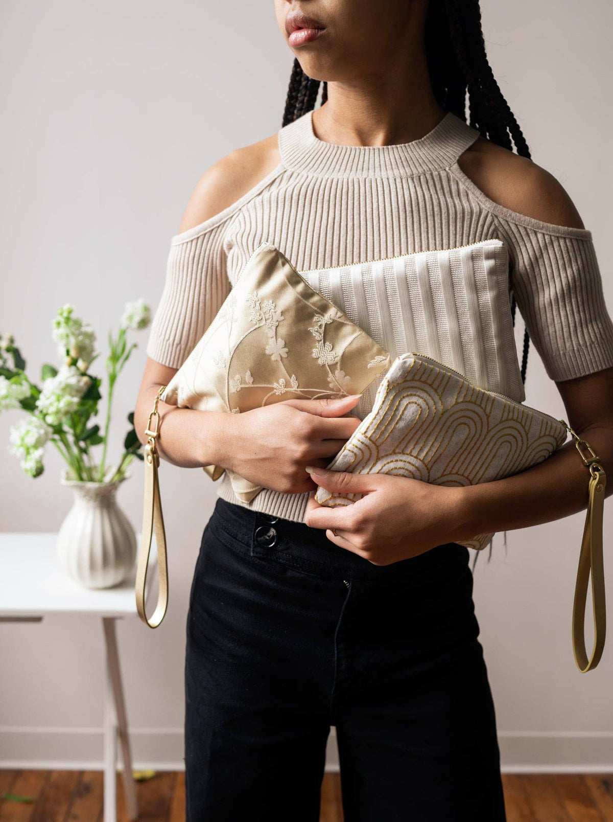A woman holding two clutch purses against her waist, a gold floral embroidered one and a Josephine Clutch with gold Art Deco arch embroidery on cream fabric