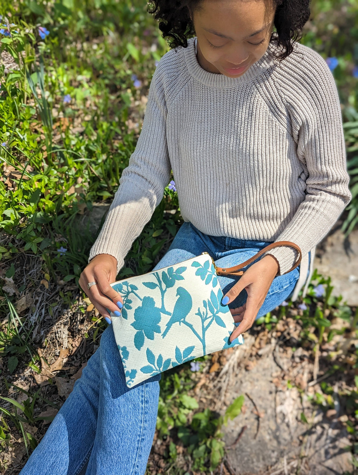 A woman holding a Bird Silhouette wristlet purse, teal bird and floral silhouette print, sitting outdoors among flowers