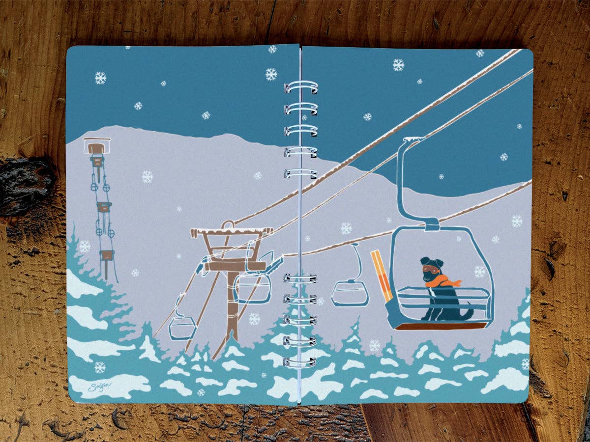 Full wraparound cover of the Ski Lift Pup spiral notebook: a pup in an orange scarf on a chairlift above snowy pines, with lift towers and falling snowflakes