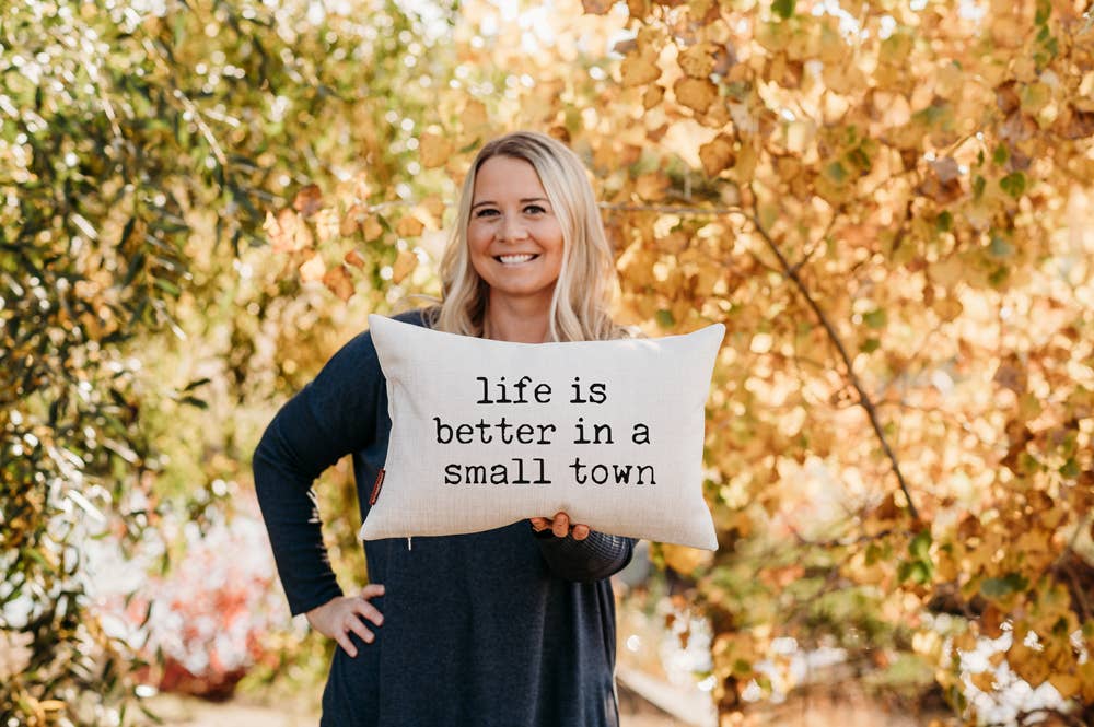 Smiling woman in a navy sweater holding the Life is Better in a Small Town Linen Pillow outdoors in front of golden fall foliage