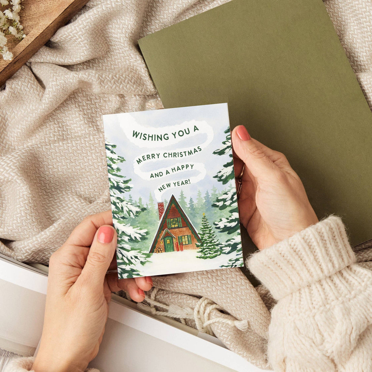 Christmas Cabin Holiday Card propped against a kraft wrapped gift tied with string on a wood table, snowy A-frame cabin design visible