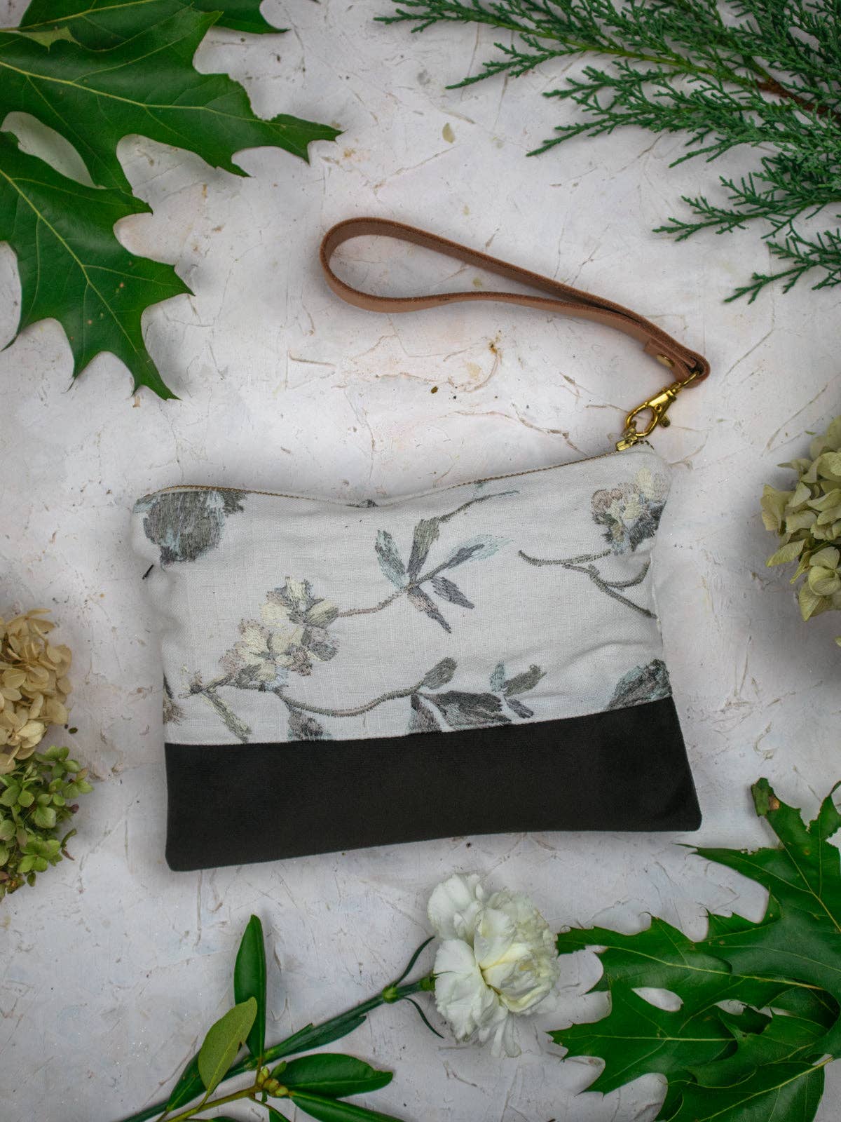 Winona Embroidered Linen wristlet, ivory linen with grey floral embroidery over a black suede base, tan leather strap, styled with leaves and a white carnation