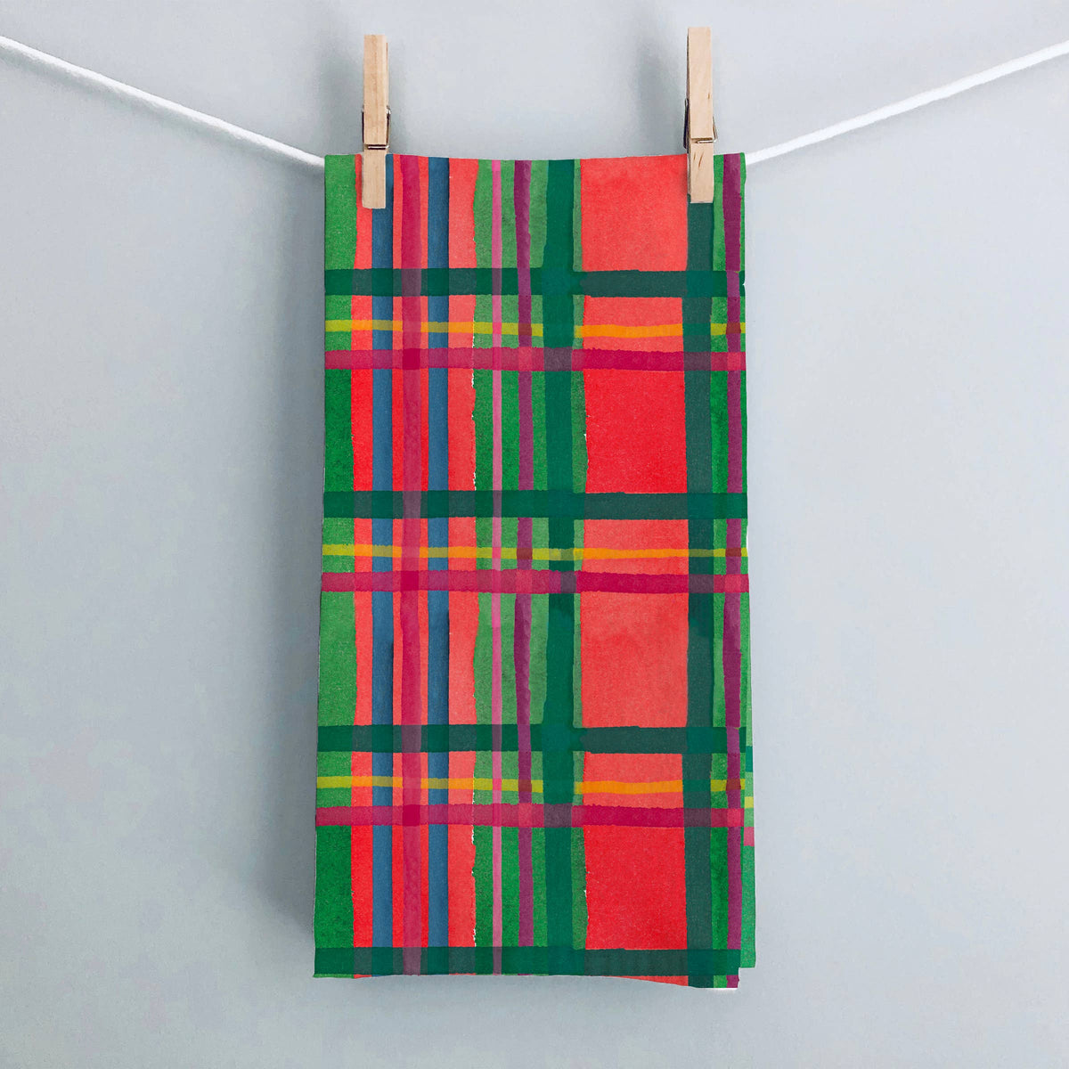 Holiday Plaid tea towel hanging from wooden clothespins on a white string against a pale gray wall