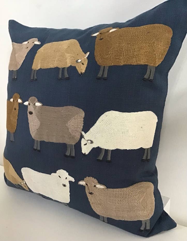 Angled view of a navy pillow embroidered with sheep in gold, taupe, white and brown, on a white surface