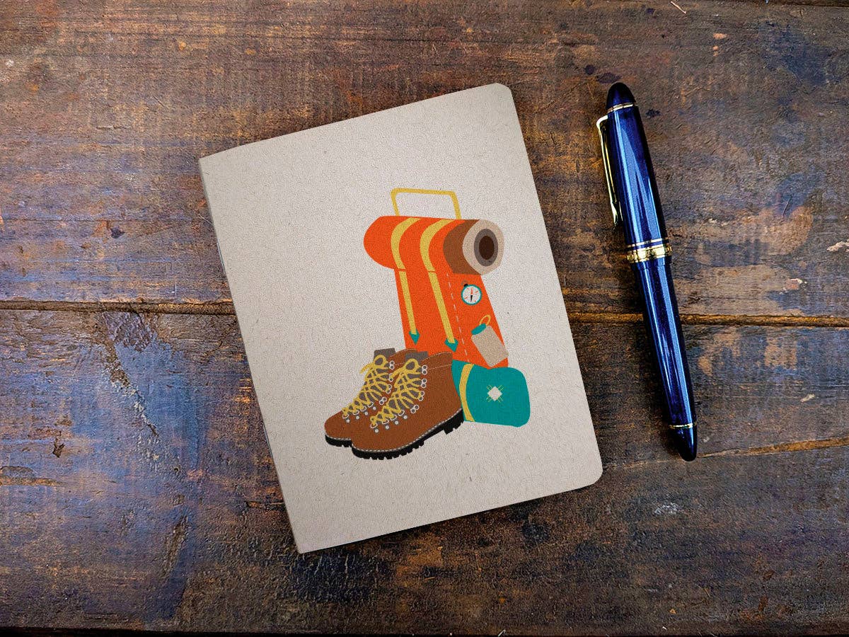Thru-Hiker pocket notebook with a kraft cover showing an orange backpack, hiking boots, and teal pouch, next to a blue fountain pen on weathered wood