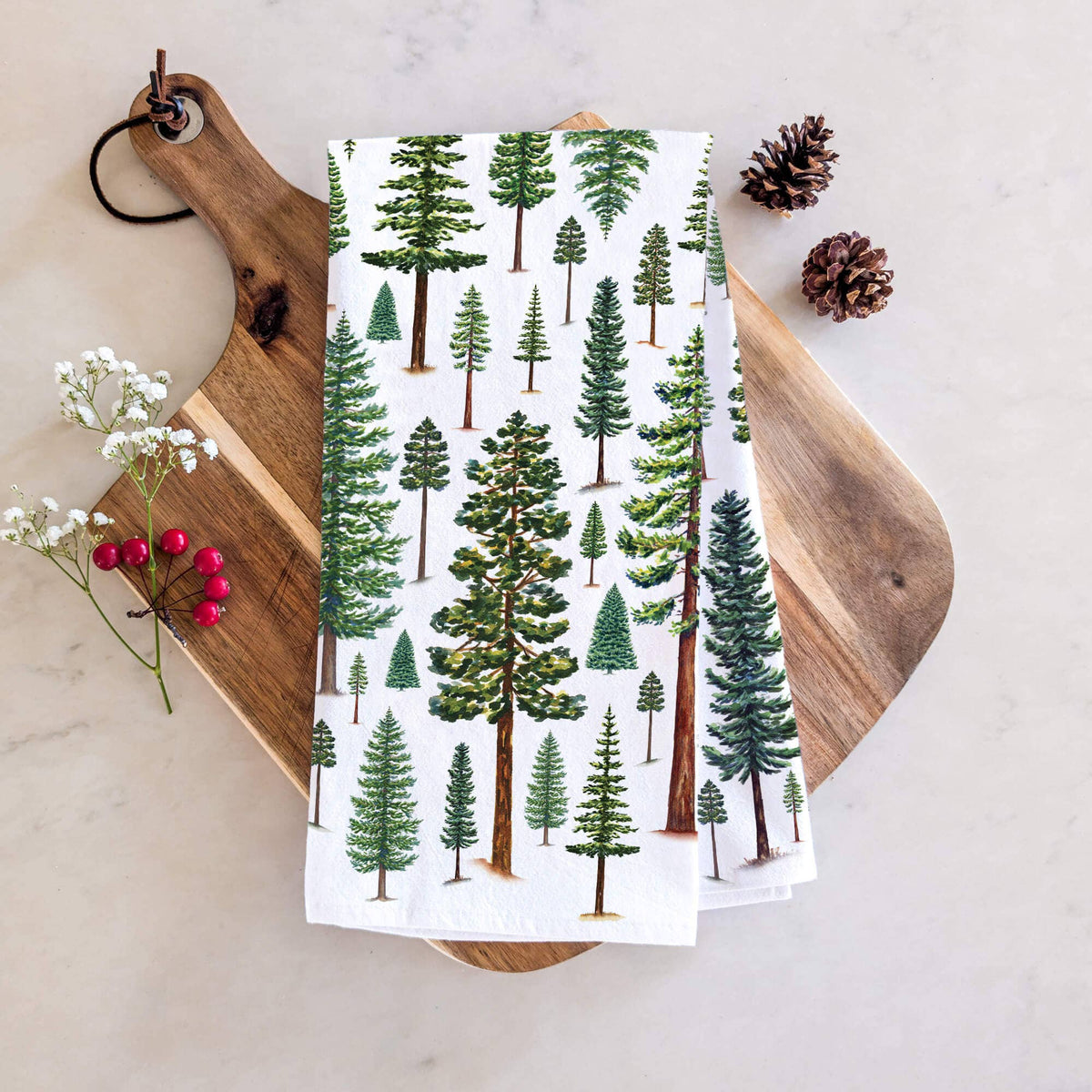 Evergreen Trees Tea Towel folded on a wood cutting board showing hand painted pine, fir, and spruce trees in shades of green, styled with pinecones and berry sprigs