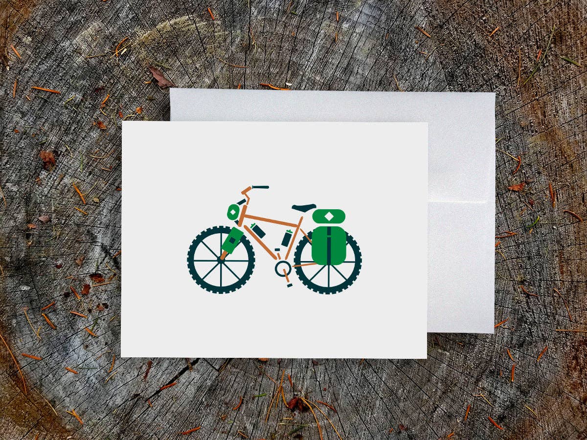 Bike-packing blank greeting card showing an orange bike loaded with green bags, on a white envelope over a weathered tree stump