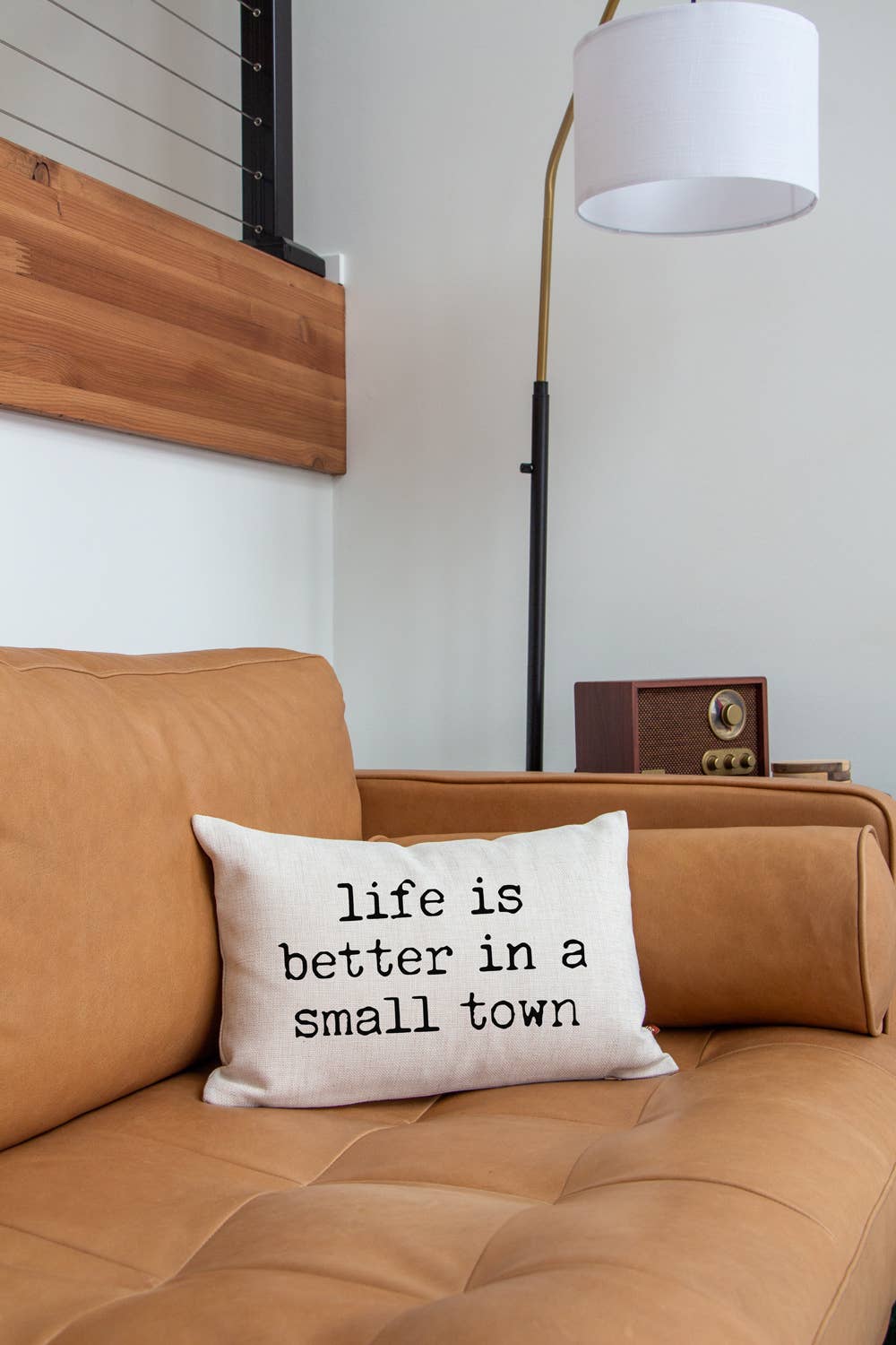 Life is Better in a Small Town Linen Pillow on a tan leather couch beneath a gold floor lamp with a white drum shade, a vintage wood radio on the armrest