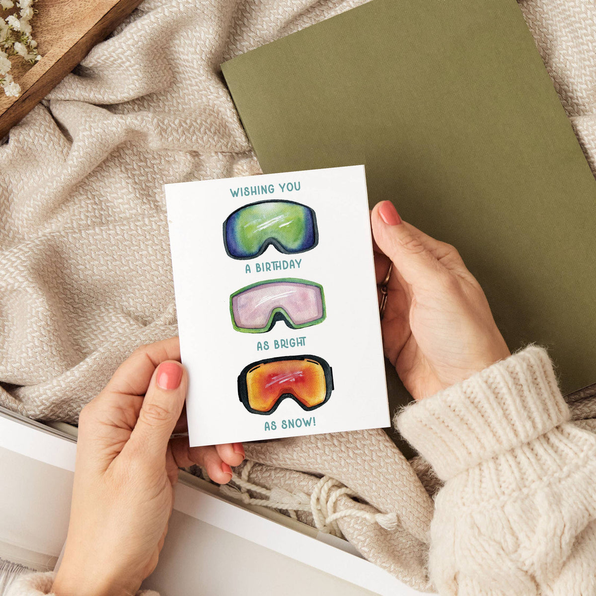 Hands holding the Bright as Snow Ski Goggles Birthday Card over a cozy scene with a knit sweater, woven blanket, and an olive green notebook