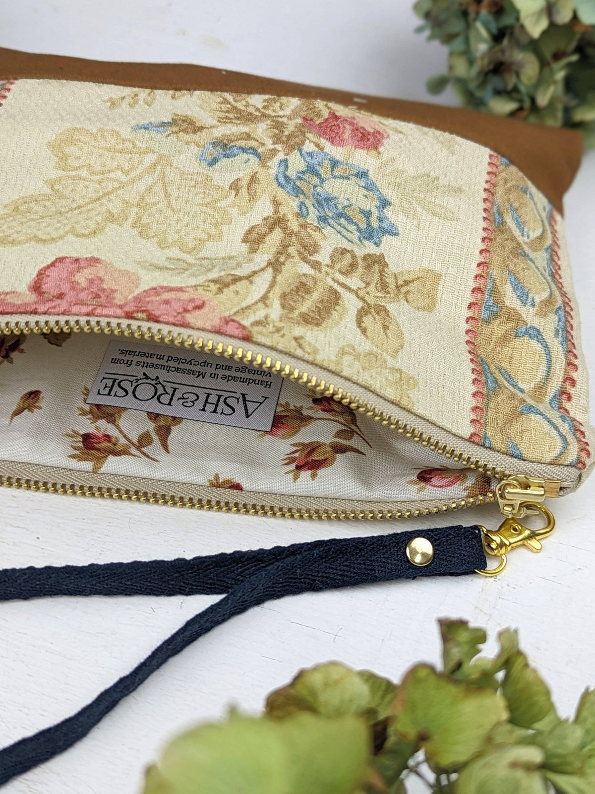 A woman holding an Antique Roses wristlet purse by its black strap, blue and pink floral fabric with tan leather bottom
