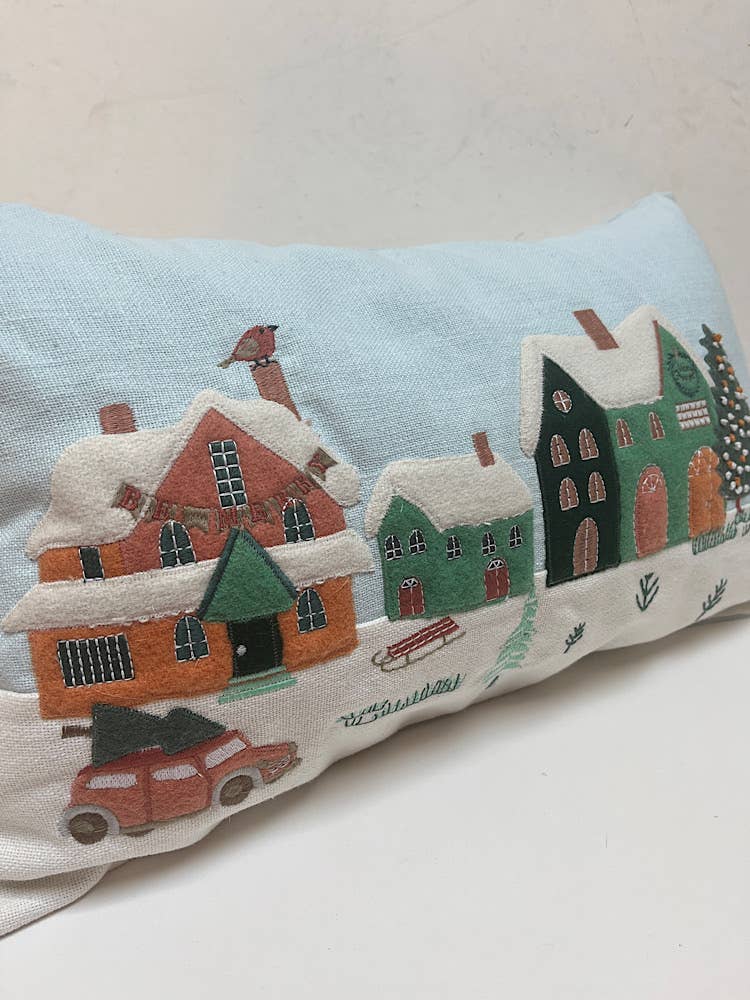 Angled view of a pale blue pillow with felt appliqued snow-covered houses, a bunting banner, a sled and a red woody car