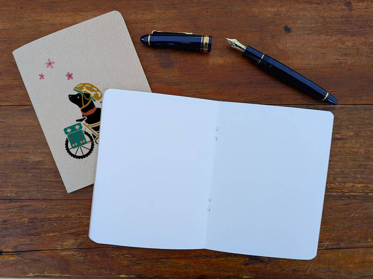 Spring Biking Pup pocket notebook open to blank white pages beside its kraft cover, with a navy fountain pen and its cap on a wood table