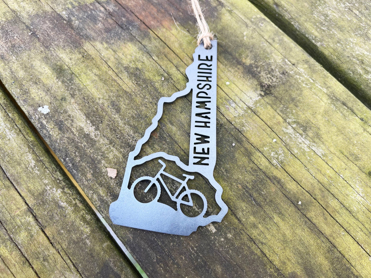 Raw steel New Hampshire mountain bike ornament with a bike cutout and NEW HAMPSHIRE lettering, flat on mossy wood boards