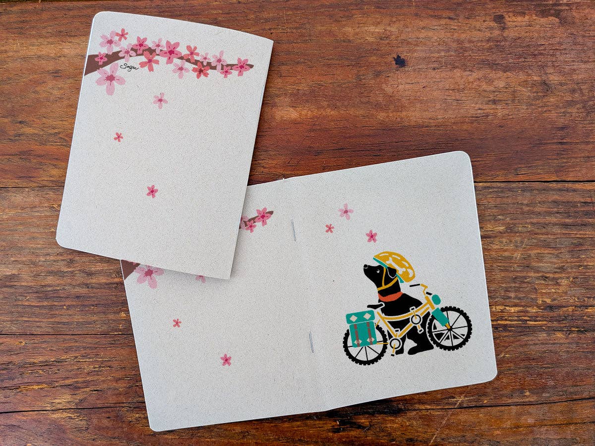 Staple-bound Spring Biking Pup notebook with a cherry blossom branch on the back cover, open beside the front cover with the dog and bike