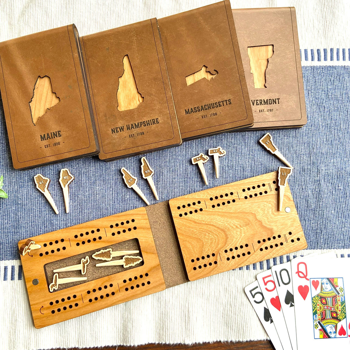 New Hampshire folding travel cribbage board open with peg storage slot, cherry wood board with peg holes and magnetic closures, next to its leather cover cut out in the shape of New Hampshire, with playing cards