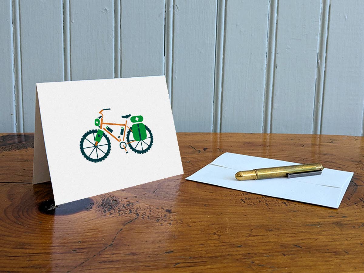 Bike-packing blank greeting card standing on a wooden table beside a white envelope and a brass pen, with a bike loaded with green bags on the cover