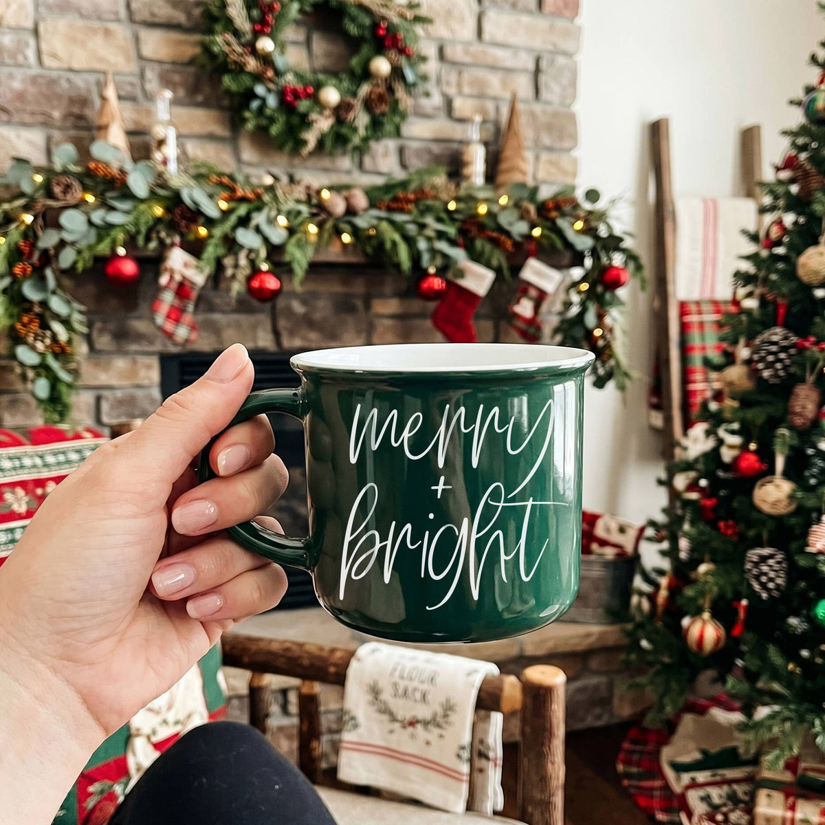Hand holding a green campfire-style ceramic mug with merry + bright in white script in front of a decorated stone fireplace, garland, stockings and Christmas tree
