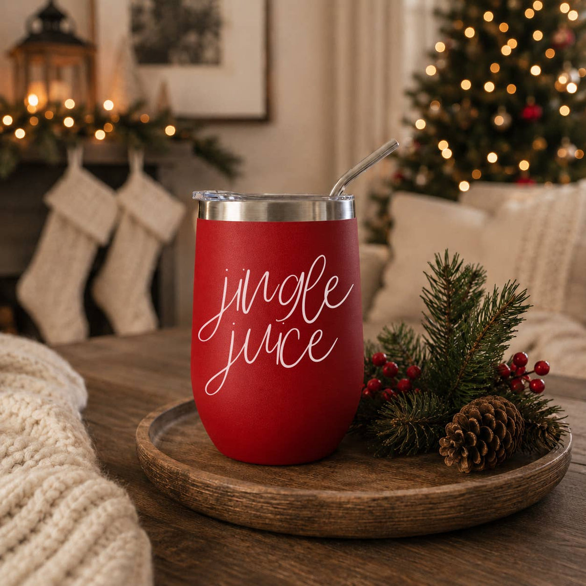 Red Jingle Juice stainless steel tumbler with a straw and clear lid on a wood tray with pine branches and a pinecone