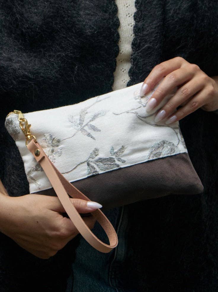 A woman holding a Winona Embroidered Linen wristlet, ivory linen with grey floral embroidery over a brown suede base, pink leather strap
