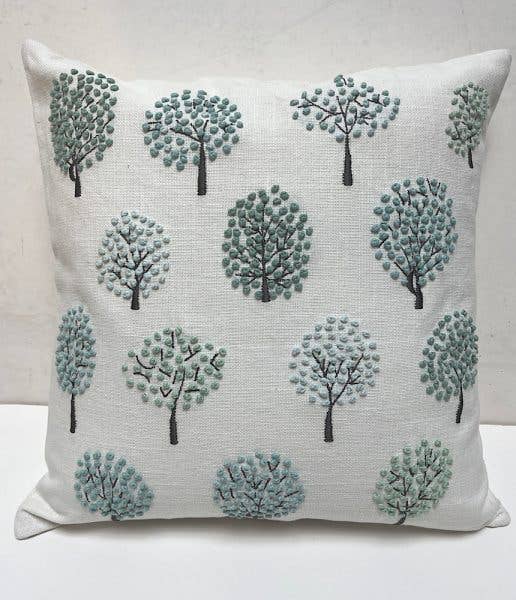 Square white cotton pillow embroidered with round-crowned trees in gray-green and blue french knots, on a white shelf