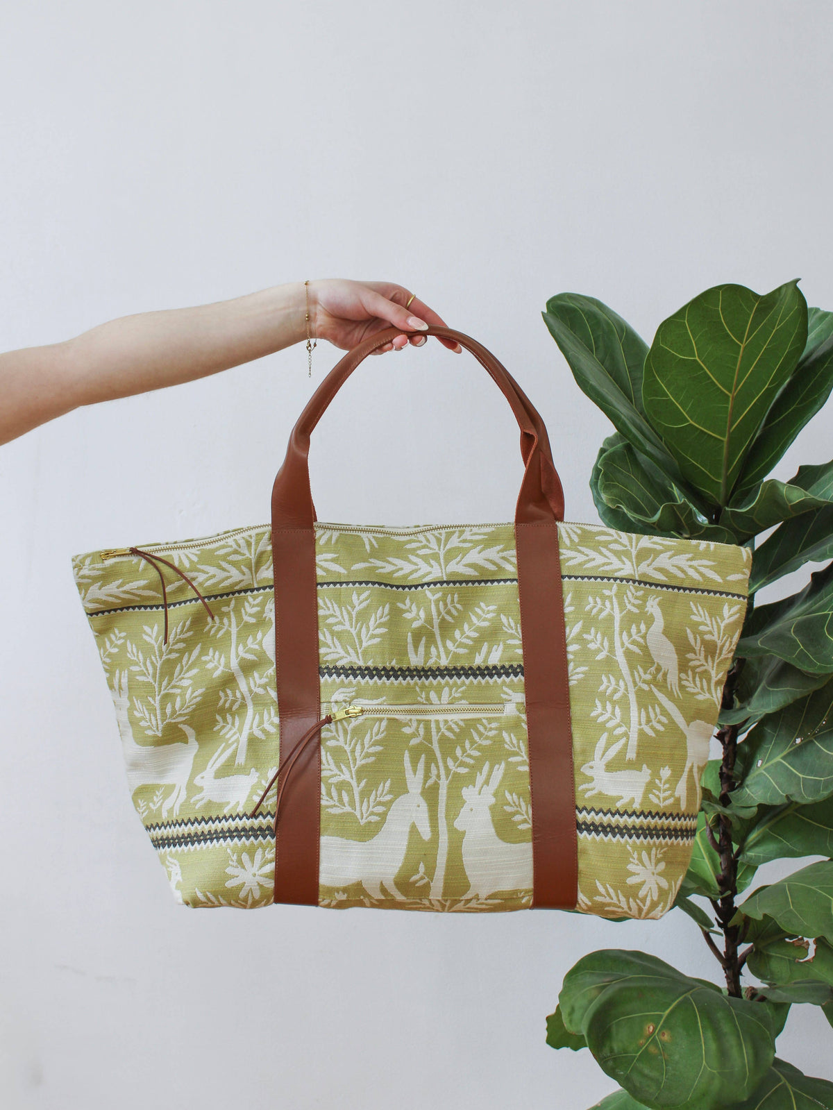A hand holding a Folk Life Carry On Travel Bag by its brown leather handles, green and cream jacquard fabric with rabbit and forest motifs
