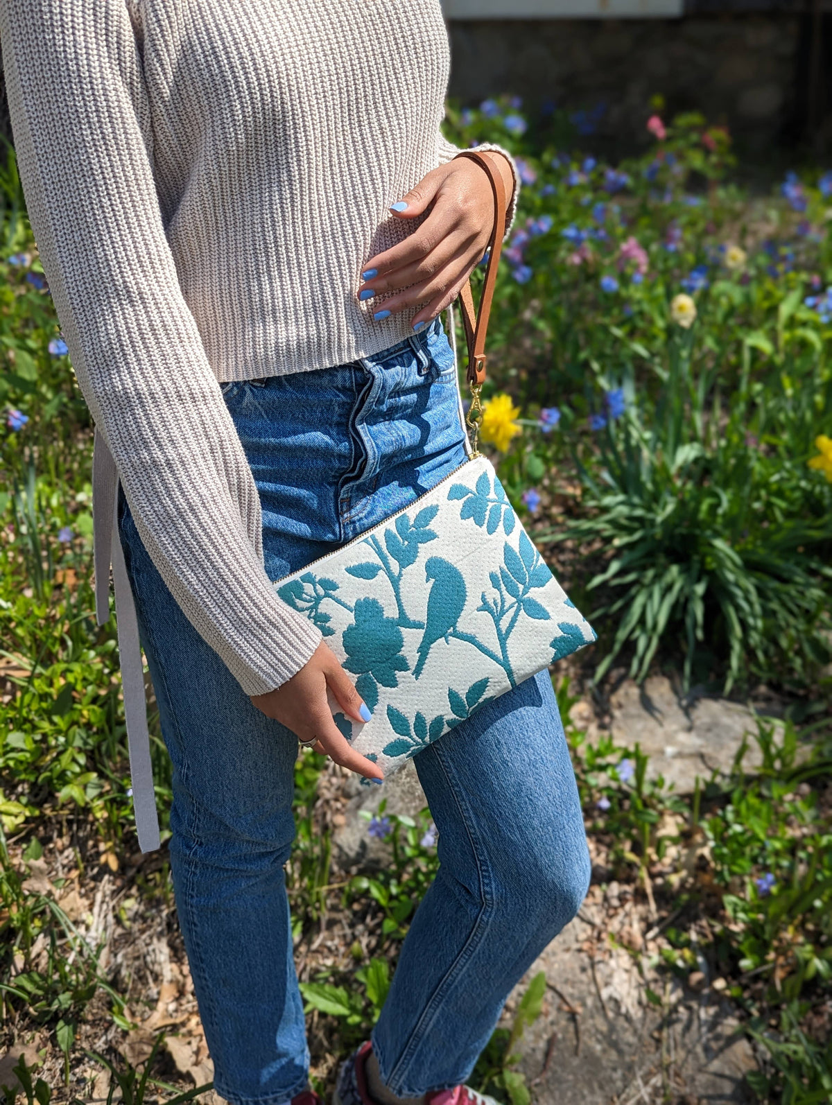 A woman holding a Bird Silhouette wristlet purse against denim jeans, teal bird and floral silhouette print, in a garden