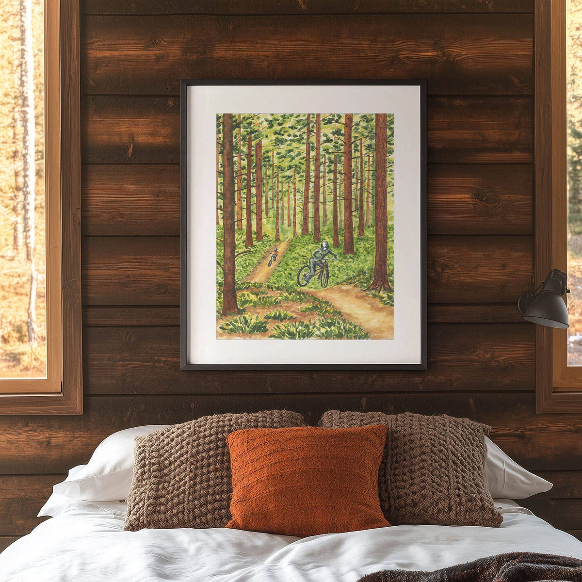 Framed Pedaling Through the Pines Watercolor Art Print in a black frame with white mat, hanging on a wood paneled wall above a bed with brown and orange pillows