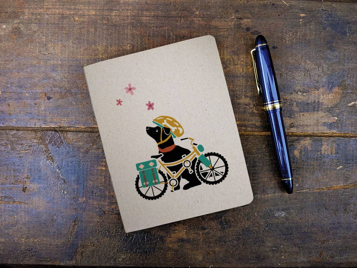 Spring Biking Pup pocket notebook with a kraft cover showing a black dog in a helmet with a bike and pink blossoms, next to a blue fountain pen