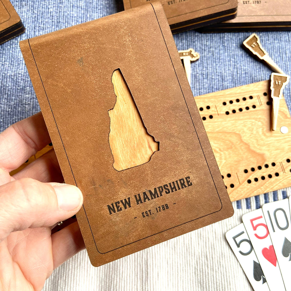 Hand holding the leather cover of the New Hampshire folding travel cribbage board, cut out in the shape of New Hampshire showing cherry wood beneath, marked New Hampshire Est. 1788