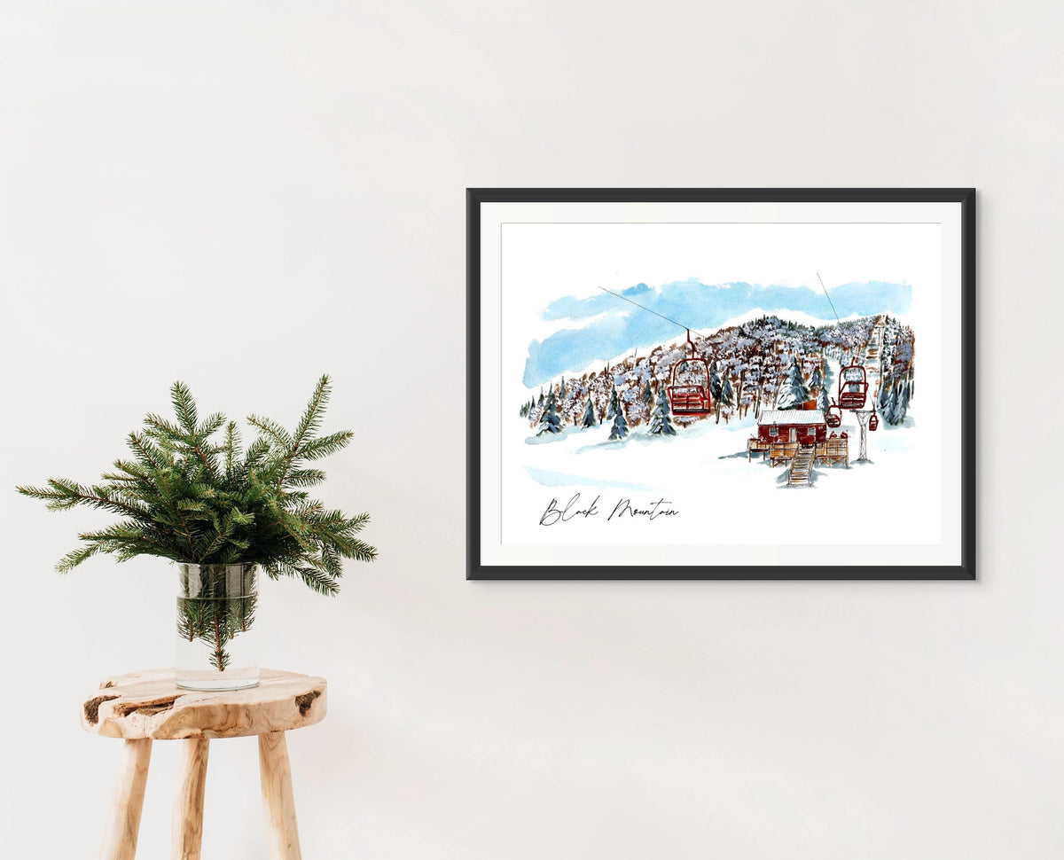 Black Mountain print in a black frame on a wall, styled with a wooden stool and a vase of pine branches