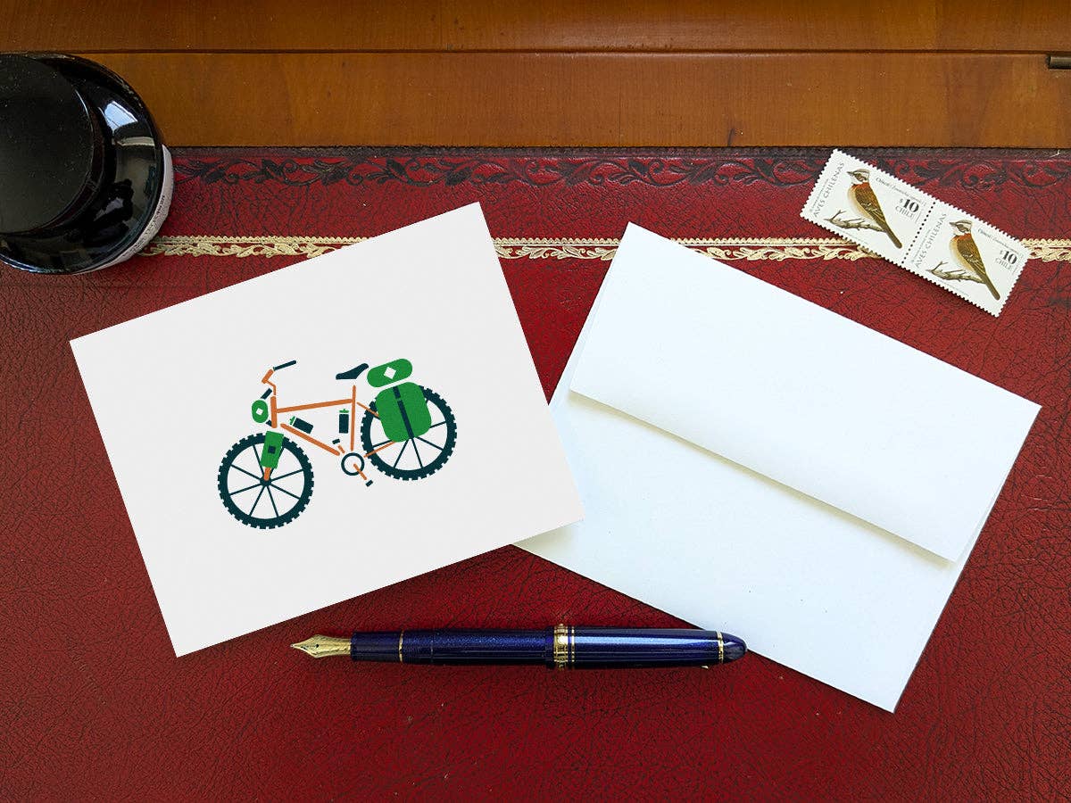 Bike-packing blank greeting card and white envelope on a red leather desk pad with an inkwell, a fountain pen, and two bird stamps