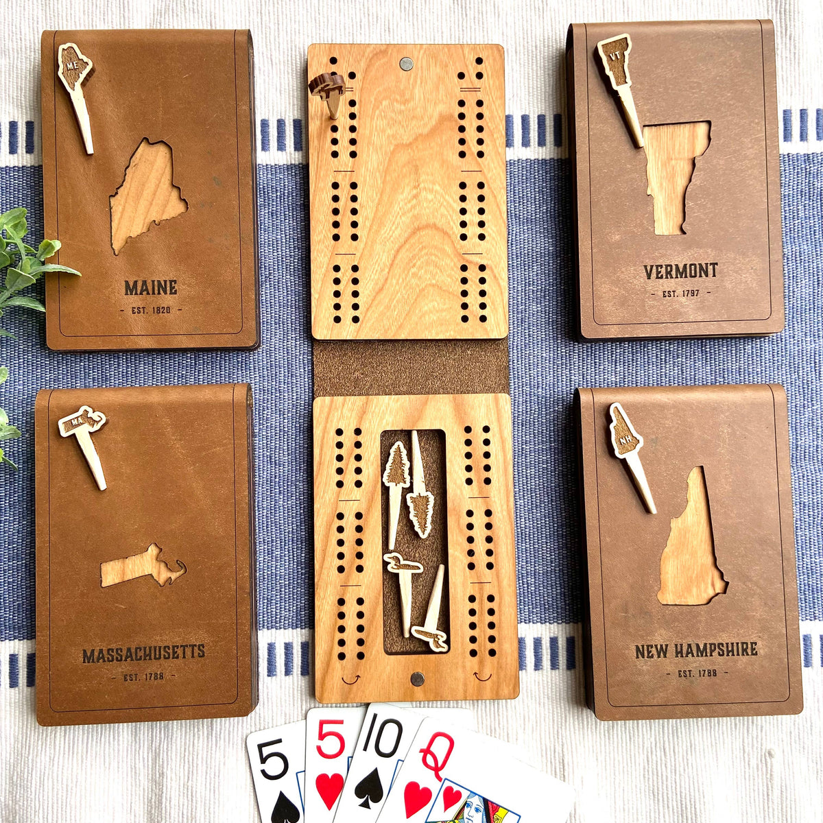 Open folding travel cribbage board showing the cherry wood peg holes and a slot holding pegs, with the New Hampshire leather cover cutout beside it and playing cards below