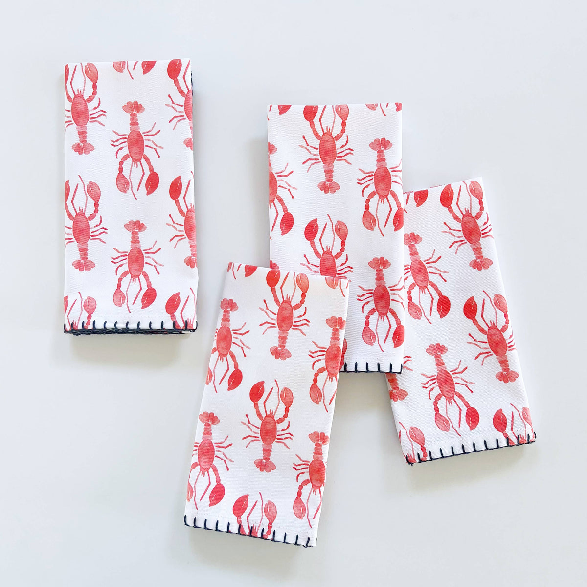 Four folded lobster napkins with red watercolor lobsters and navy whipstitch edges on a white surface