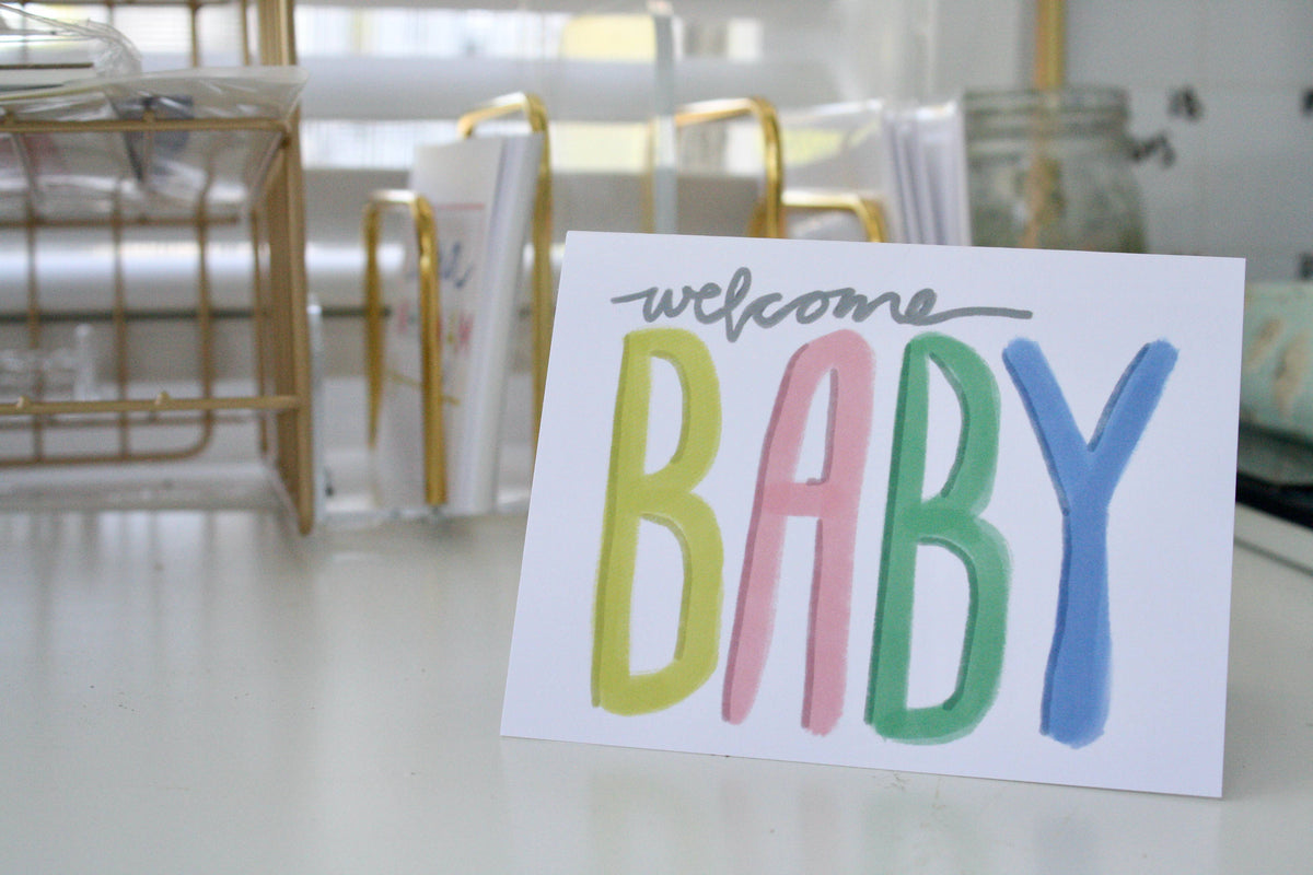 Welcome Baby Greeting Card propped on a desk near gold wire organizers and a glass jar, colorful yellow pink green and blue lettering spelling BABY