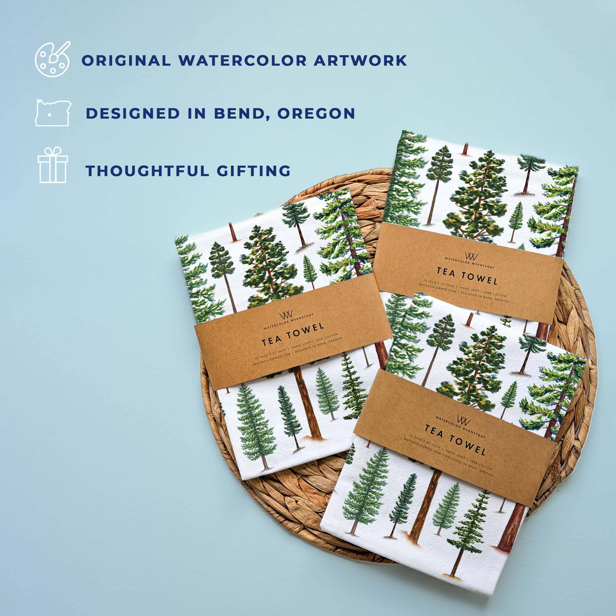 Original watercolor artwork, designed in Bend Oregon, and thoughtful gifting icons next to several folded Evergreen Trees Tea Towels wrapped in kraft Watercolor Wednesday tea towel bands on a woven mat