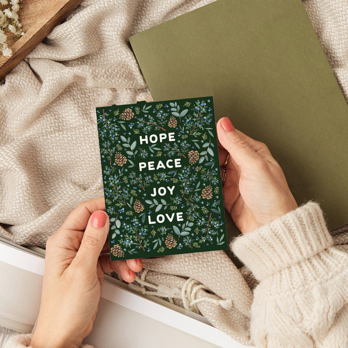 Hands holding the Winter Greenery Holiday Card over a cozy scene with a knit blanket and an olive green notebook