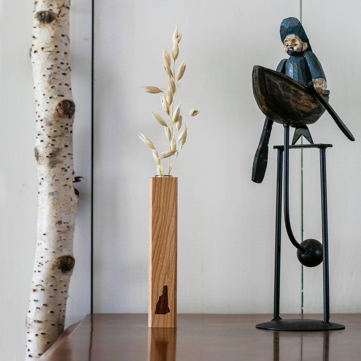 New Hampshire Bud Vase in the Wood finish holding a dried wheat stem, styled on a wood table next to a birch branch and a wooden bird figurine on a stand