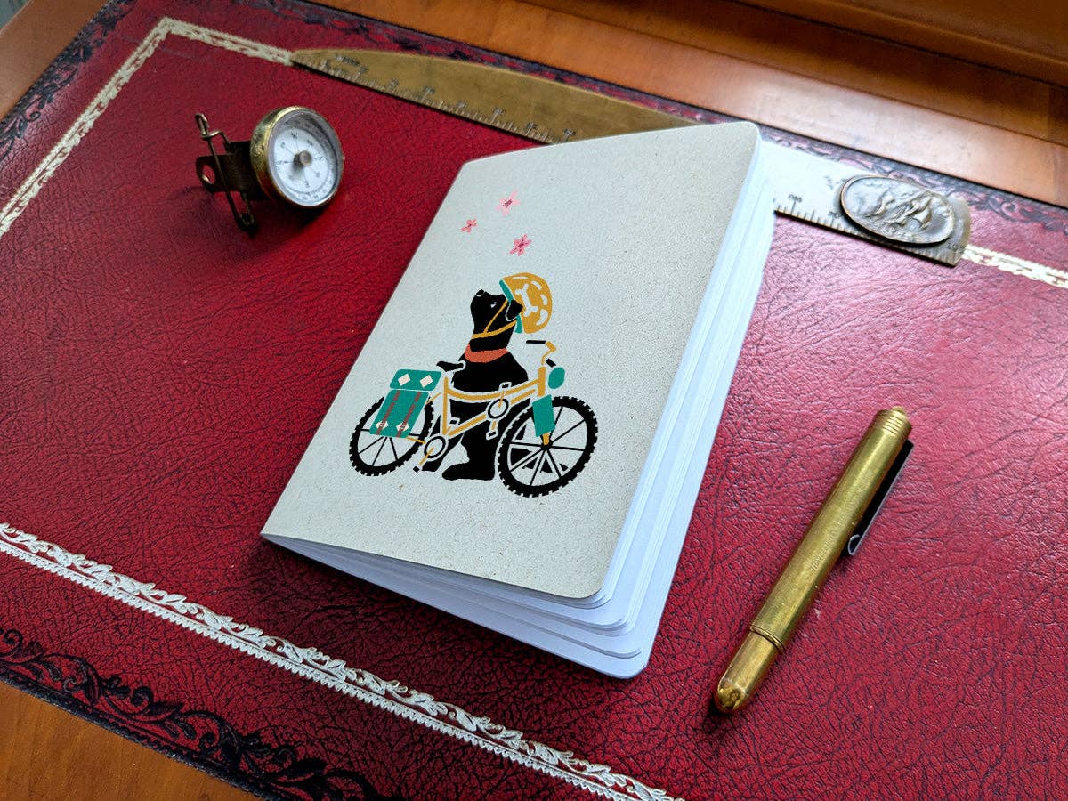 Spring Biking Pup pocket notebook with a black dog in a yellow helmet beside a bike with teal panniers, looking up at pink blossoms, on a red leather desk pad
