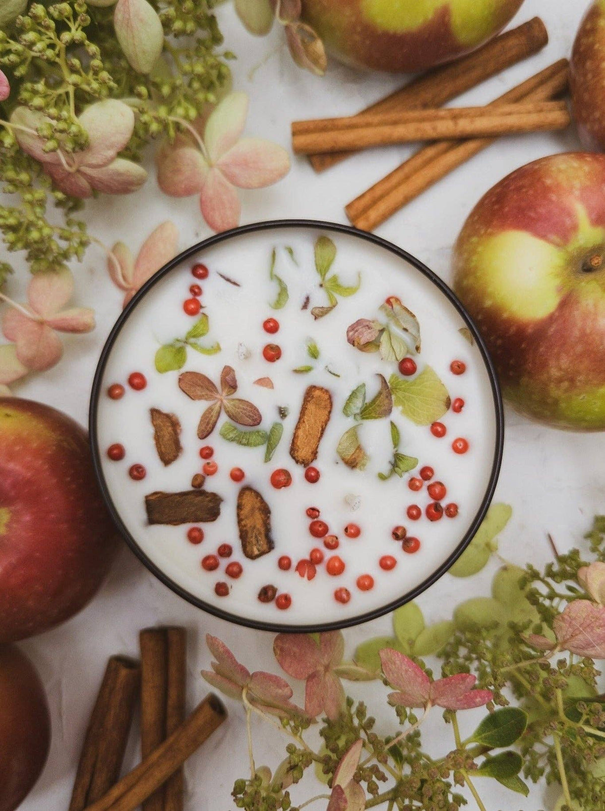 HARVEST Apple Spice soy candle, wax topped with cinnamon bark pieces, dried hydrangea petals, and pink peppercorns, surrounded by apples and cinnamon sticks